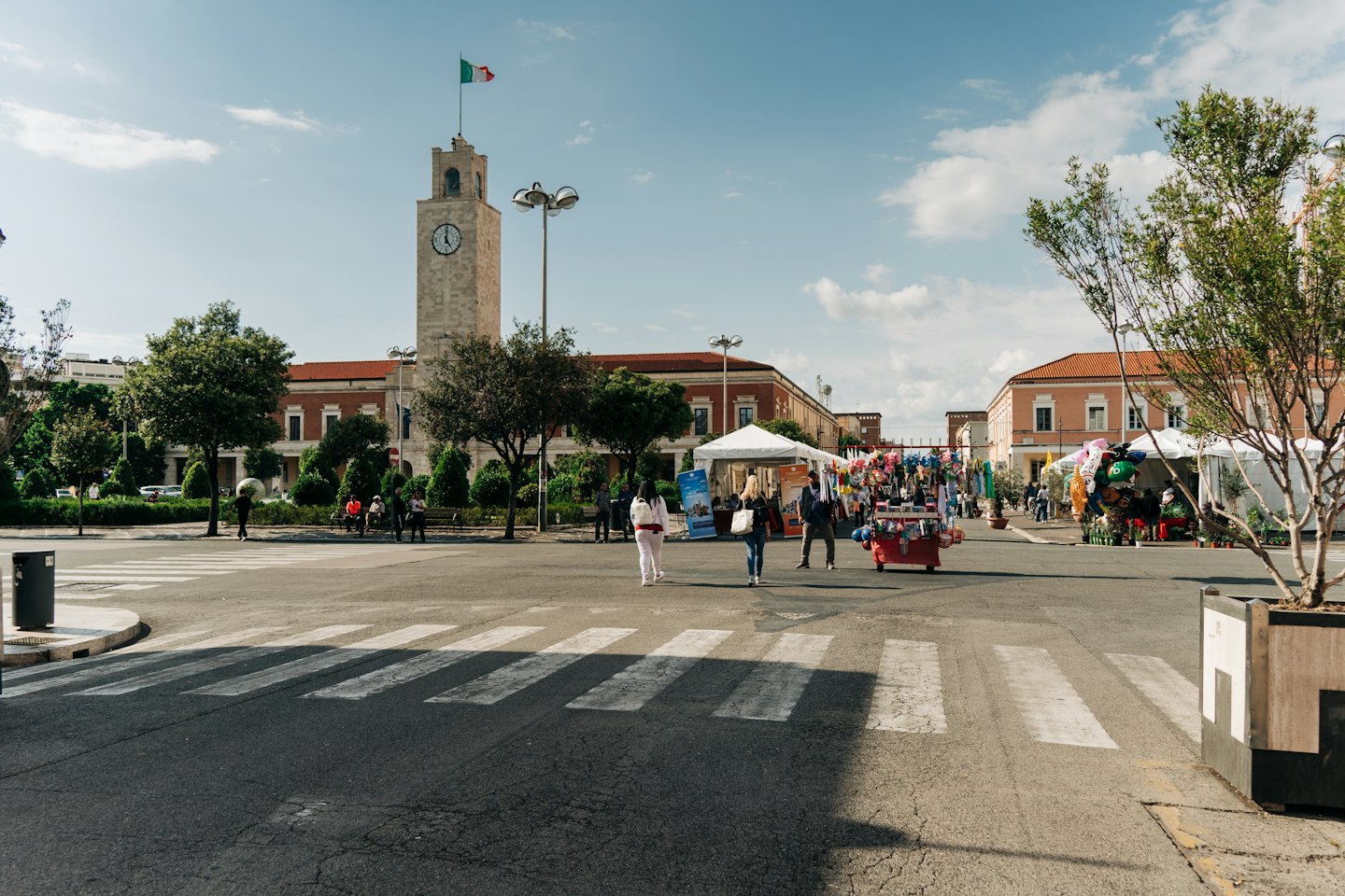 Piazza del paese con torre dell'orologio, bancarelle e persone.