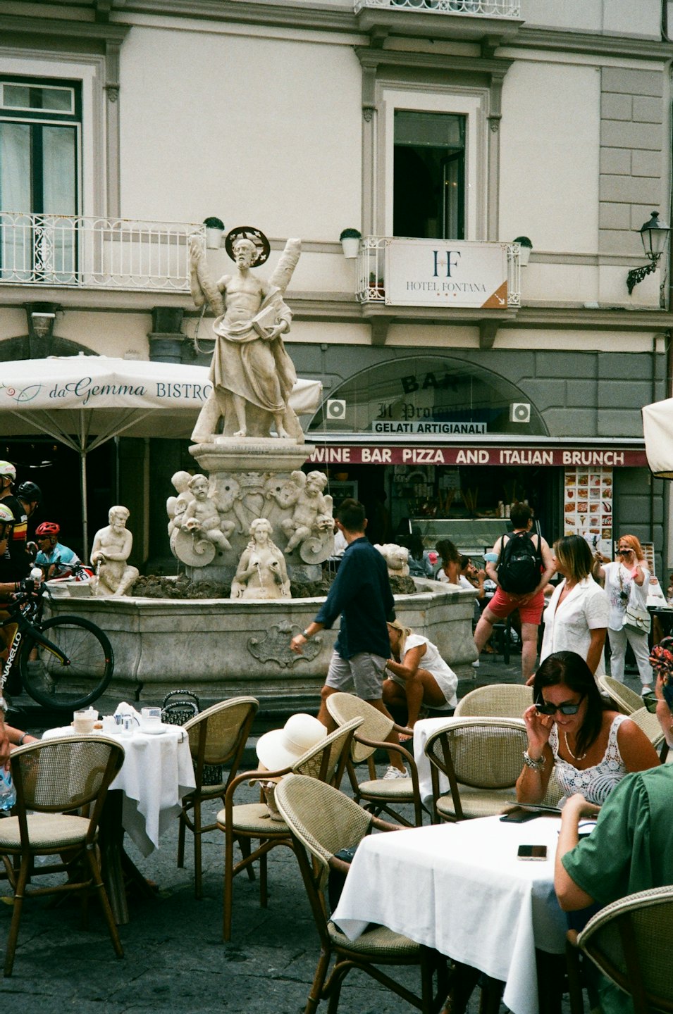 Persone cenano a tavoli all'aperto vicino a una grande fontana di pietra.